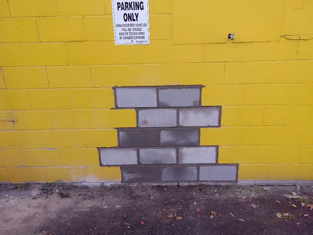 This image shows a yellow brick wall with a Parking Only sign posted on it The sign warns that unauthorized vehicles will be towed away at the owners expense The wall also has a partially damaged section where some of the bricks have been removed creating a small gap in the wall